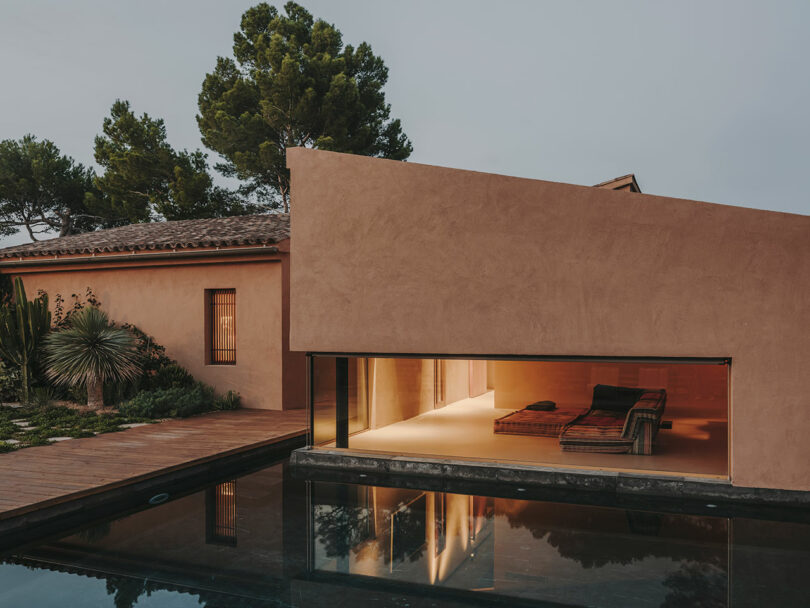 Modern house with a flat, angular roof, large glass windows, and an illuminated interior lounge area overlooking a reflective pool, surrounded by trees and plants.