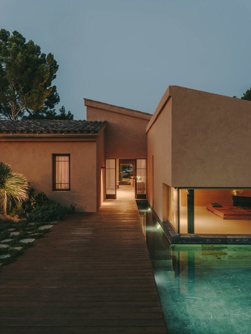 Modern house with warm lighting, wood walkway, and adjacent reflecting pool at dusk, surrounded by trees and landscaped greenery.