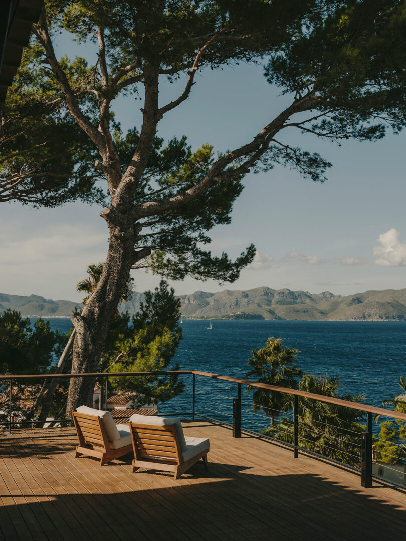 Two wooden lounge chairs on a deck overlook the sea, with tall trees nearby and mountains visible in the distance under a clear sky.