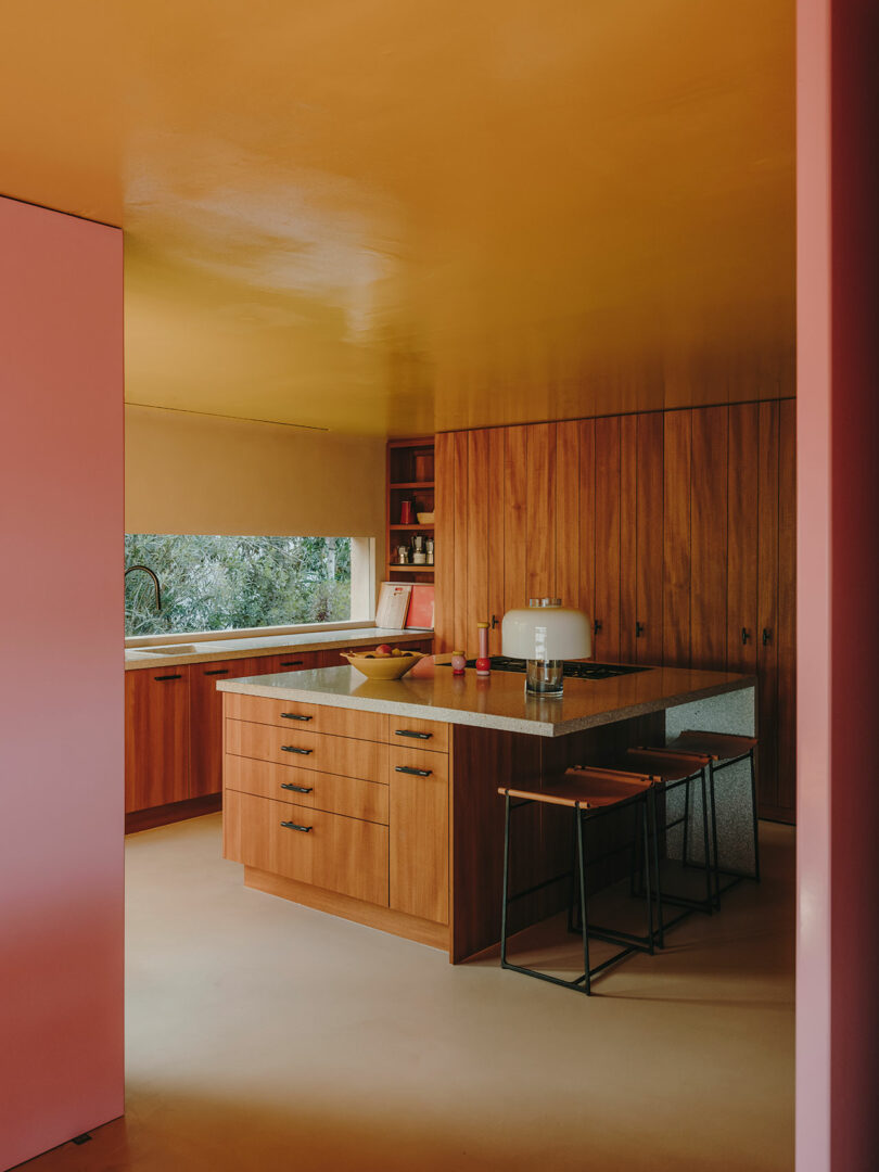 Modern kitchen with wooden cabinets, island with stools, pink and yellow walls and ceiling, countertop appliances, and a window overlooking greenery.