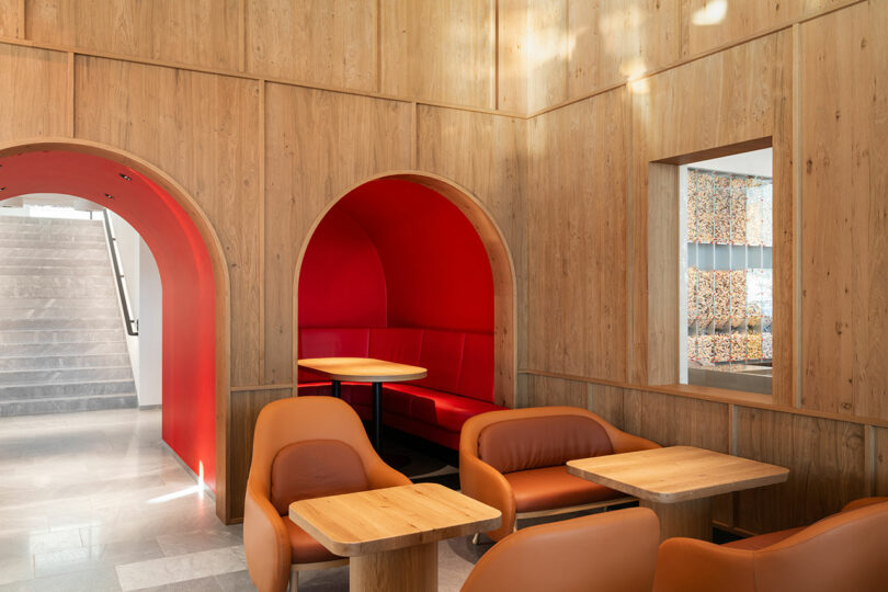 A modern seating area with brown chairs and wooden tables in front of arched wooden wall alcoves, one with red upholstery and a table, near a staircase.