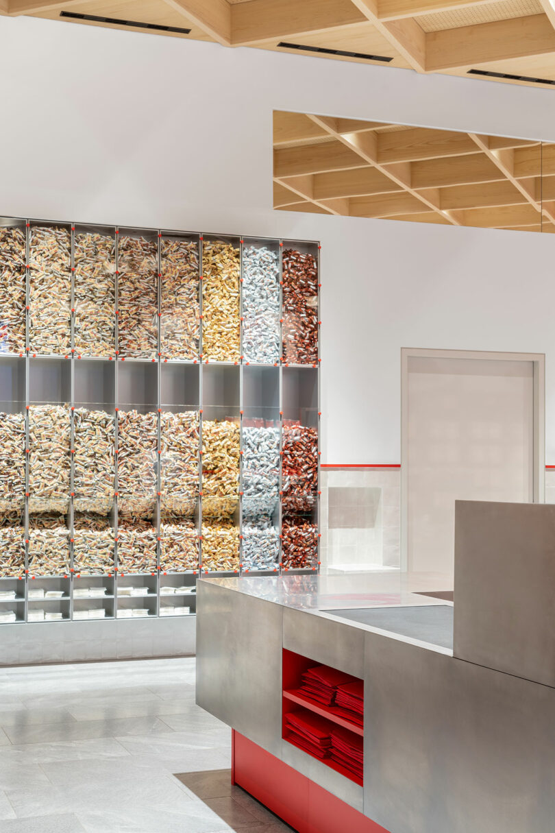 A modern store interior with a wall of organized clear bins containing various dried pasta shapes, a metal counter, and a light wood ceiling.