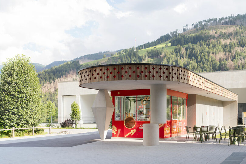A small modern building with a geometric roof design and red glass walls stands outside, with green chairs nearby and mountains in the background.