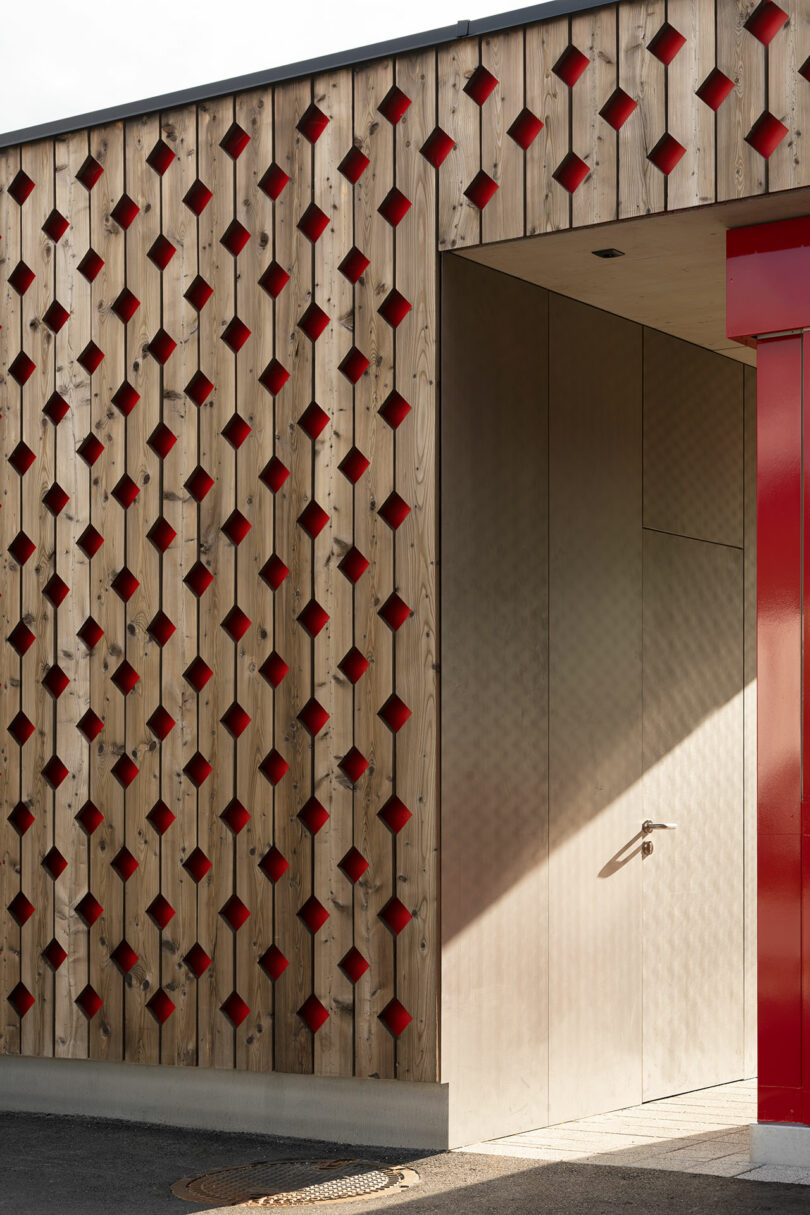 Wooden facade with a geometric pattern of diamond-shaped cutouts backed by red, next to a modern entrance with a red frame and a door casting a shadow.