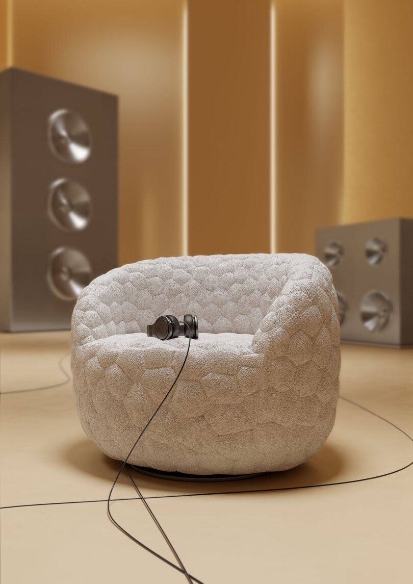 A round, textured armchair sits on a beige floor with headphones resting on the seat; large silver speakers stand in the background.
