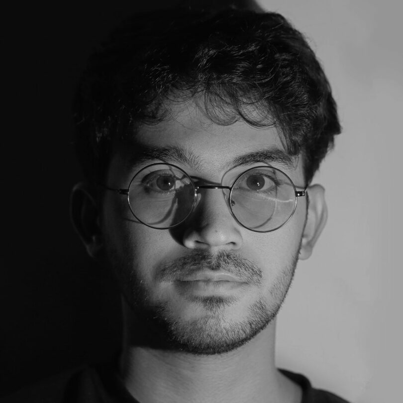 A young man with short hair, glasses, and facial hair looks directly at the camera. The lighting creates a stark contrast between light and shadow on his face.