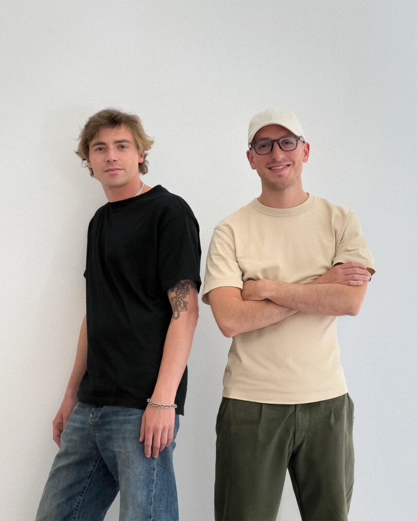 Two people stand side by side against a plain white wall; one wears a black shirt and jeans, the other wears a beige shirt, dark pants, a white cap, and glasses.