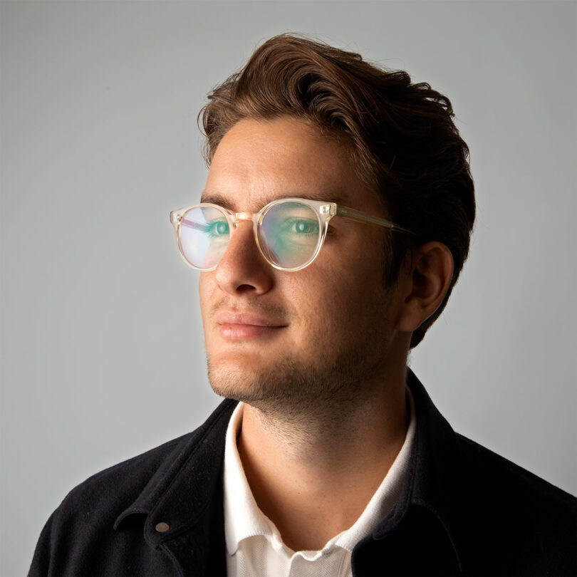 A young man with brown hair and glasses, wearing a white shirt and dark jacket, looks slightly upward against a plain background.