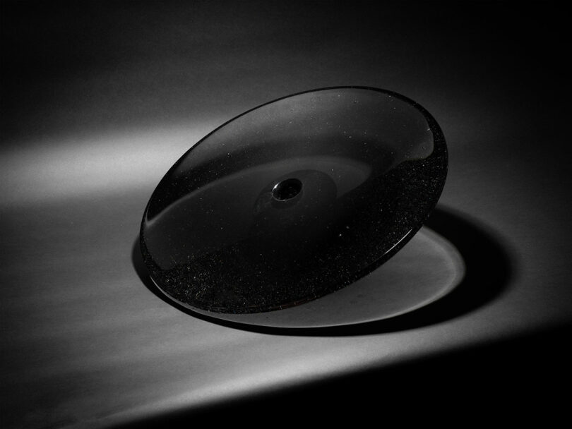 A transparent, shallow glass disc by Claesson Koivisto Rune with a small central indentation is lit from the side, casting a shadow on a smooth, grey surface.