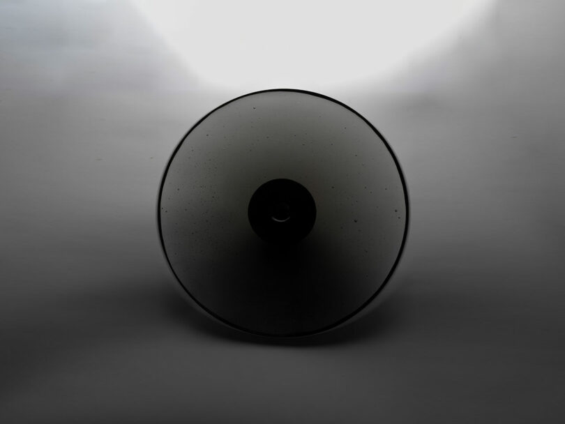 A round, dark translucent object, reminiscent of Claesson Koivisto Rune's minimalist style, is centered against a grey background, with a gradient halo of light behind it.