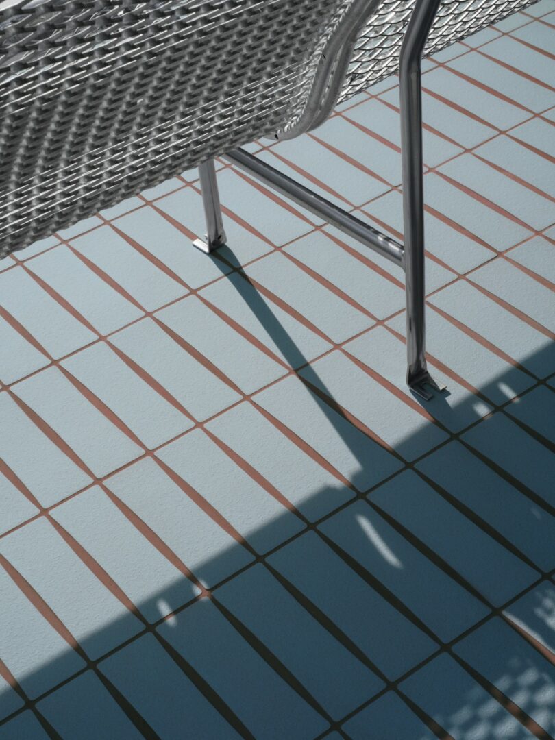Metal chair casting a shadow on a tiled floor with blue and brown rectangular tiles, partially shaded by another object