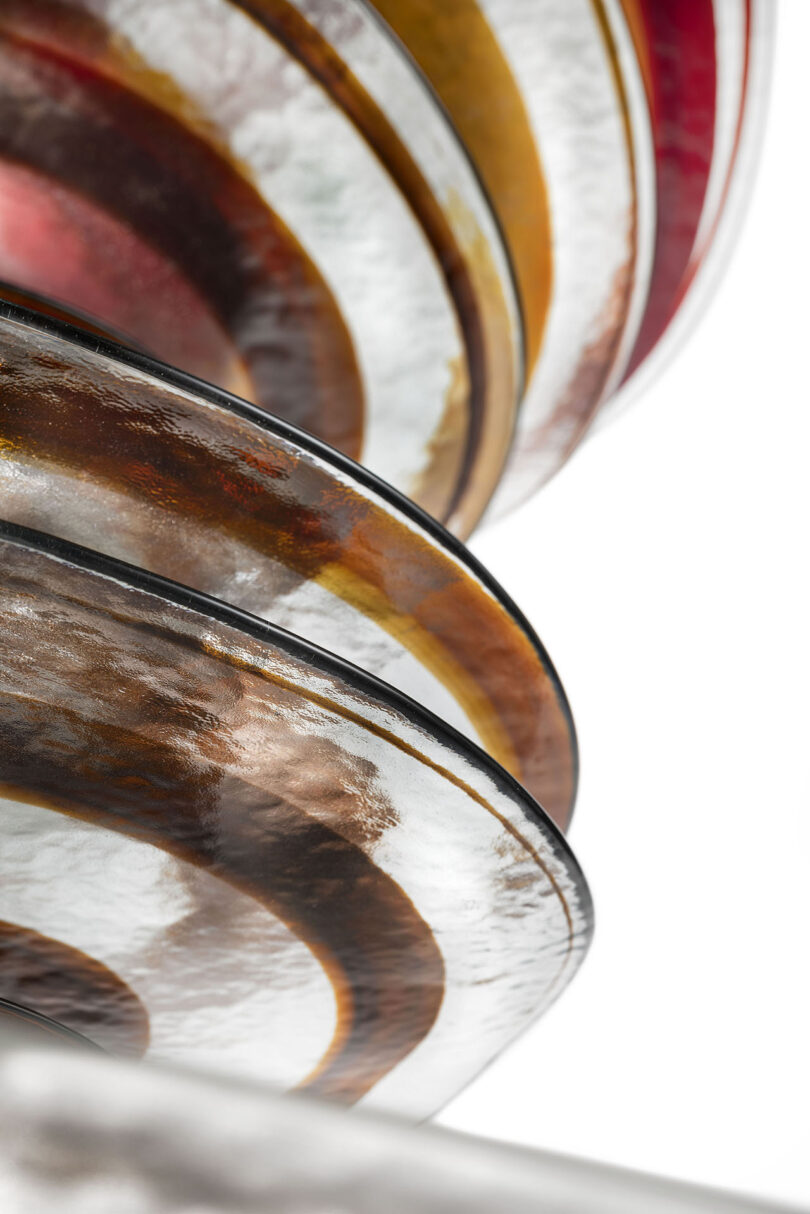 Close-up of stacked glass plates with textured surfaces and swirling patterns in brown, amber, and red tones against a white background.