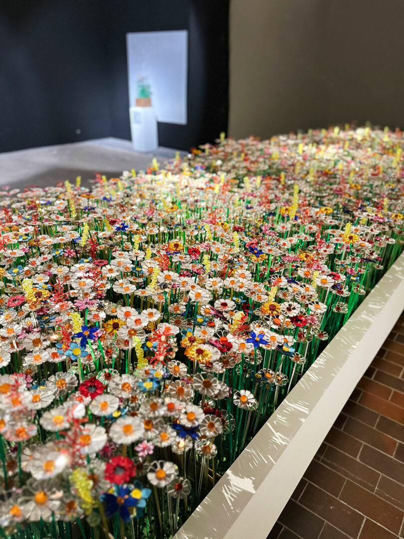 A large installation of colorful artificial flowers, reflecting diversiveness, is arranged densely in a rectangular bed inside a modern indoor space with dark walls.