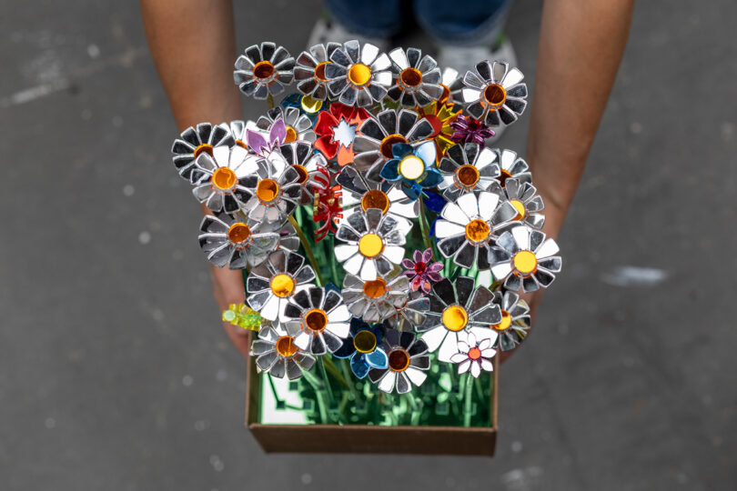 A person holds a box filled with artificial flowers made from shiny metal and colored materials, arranged to resemble daisies.