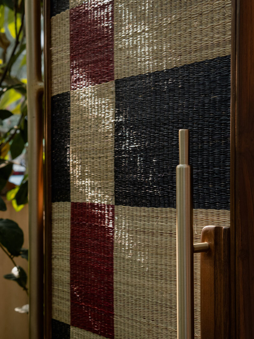 A close-up of a woven door with a red, black, and tan checkered pattern, featuring a vertical metal handle and partial view of a plant on the left.