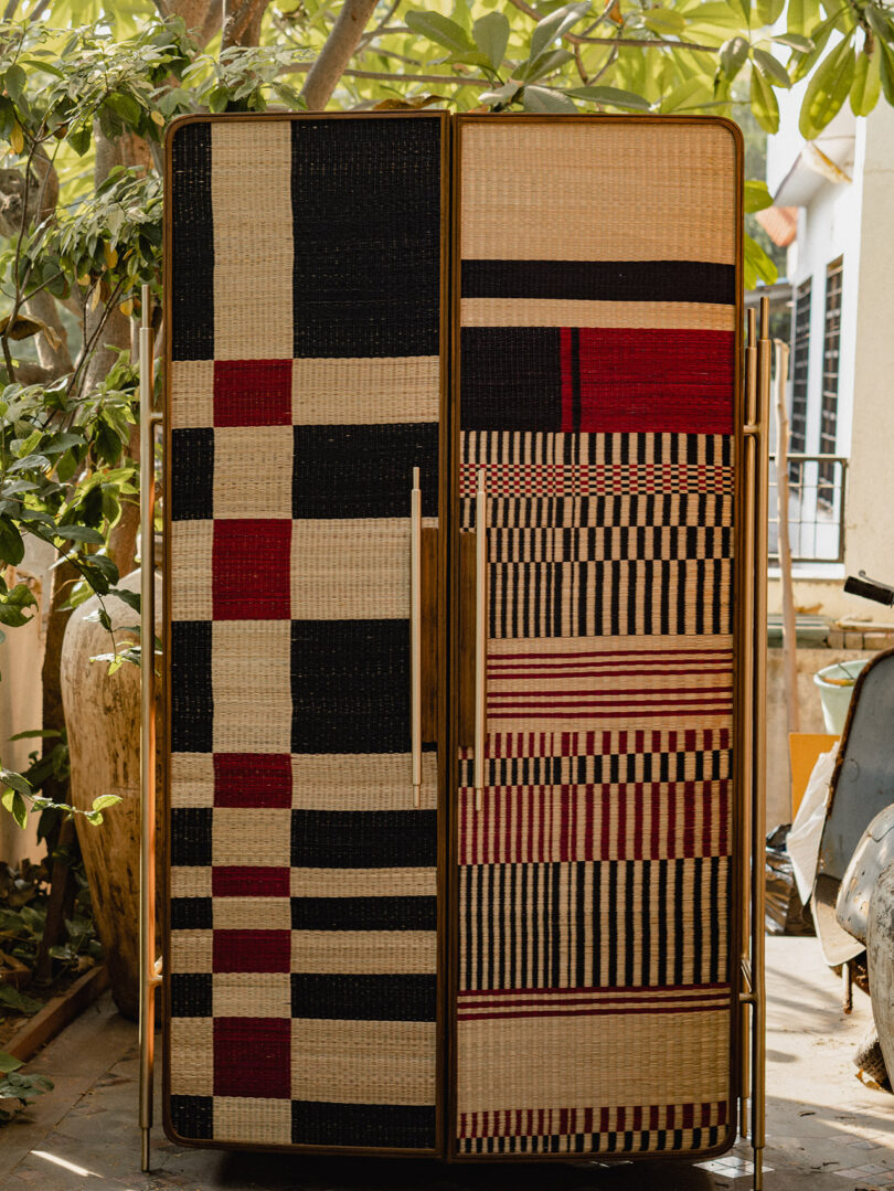 A woven cabinet with geometric patterns in black, red, and beige stands outdoors near potted plants and building walls.