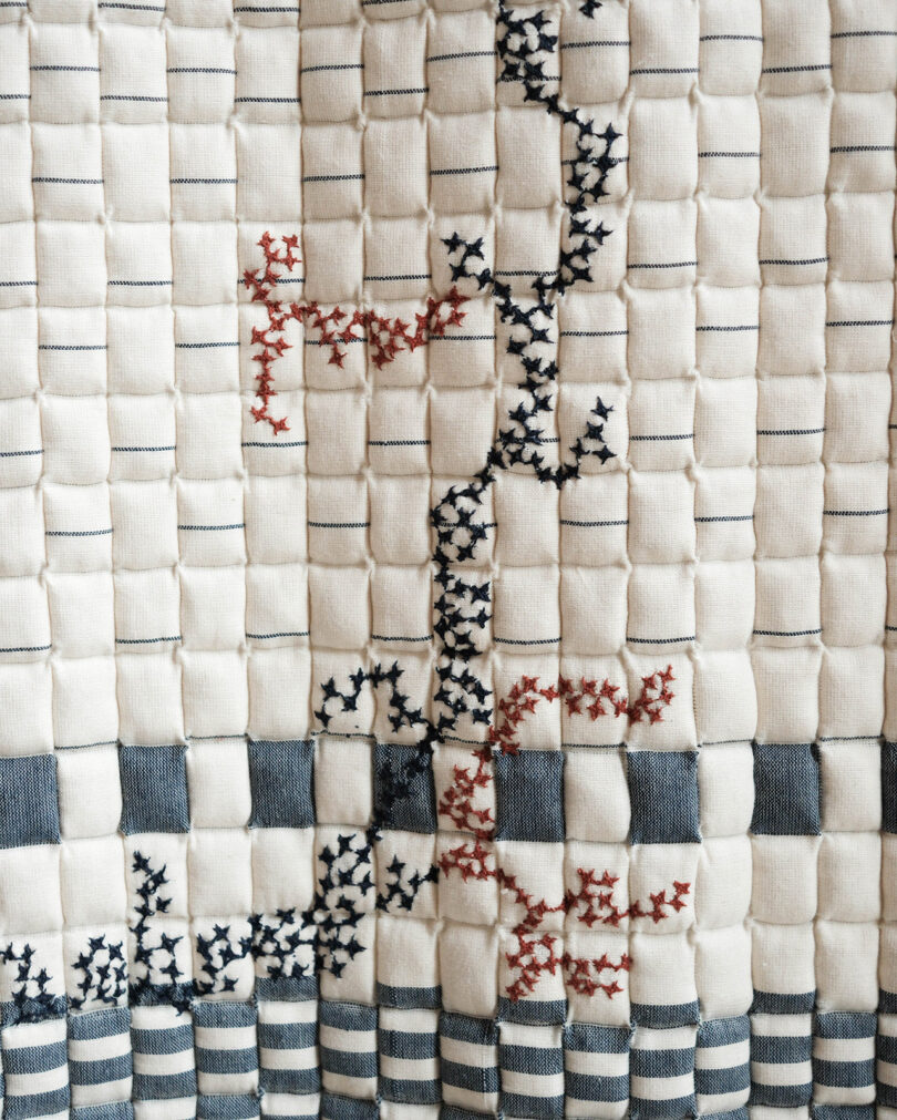 A quilted textile with vertical and horizontal stitched lines, decorated with black and red cross-stitch patterns forming abstract shapes.