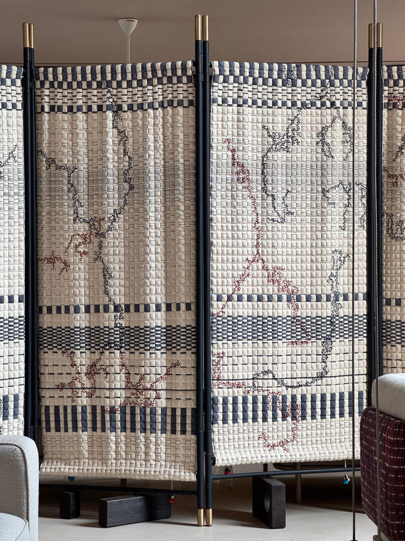 A fabric room divider with a woven, checkerboard pattern in white and blue, supported by black vertical poles, stands between modern furniture in a contemporary interior.