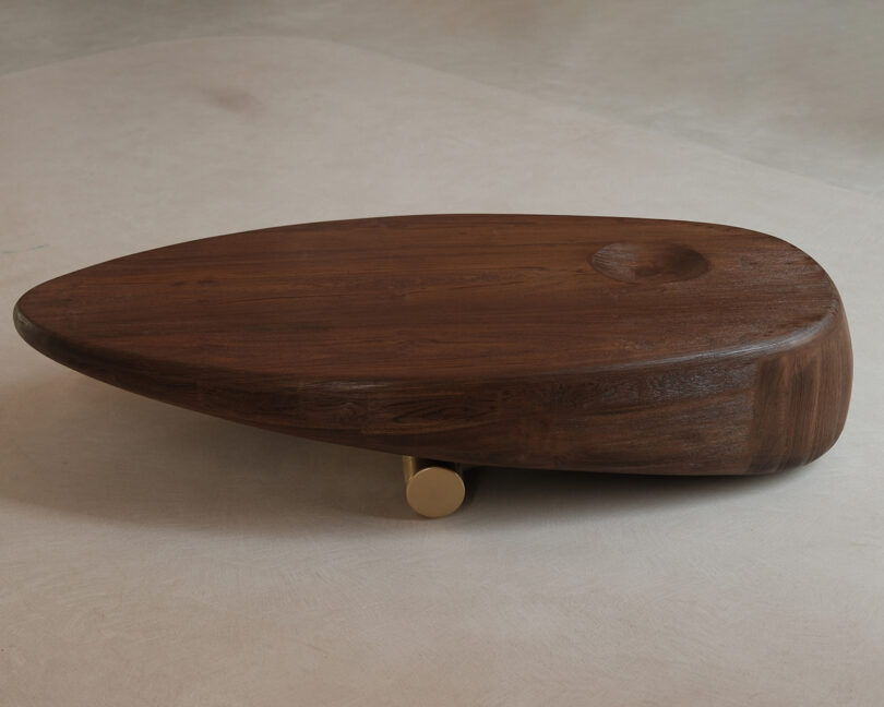 Low, oval-shaped wooden coffee table with a smooth finish, rounded edges, a small circular indentation, and a single gold cylindrical leg underneath.