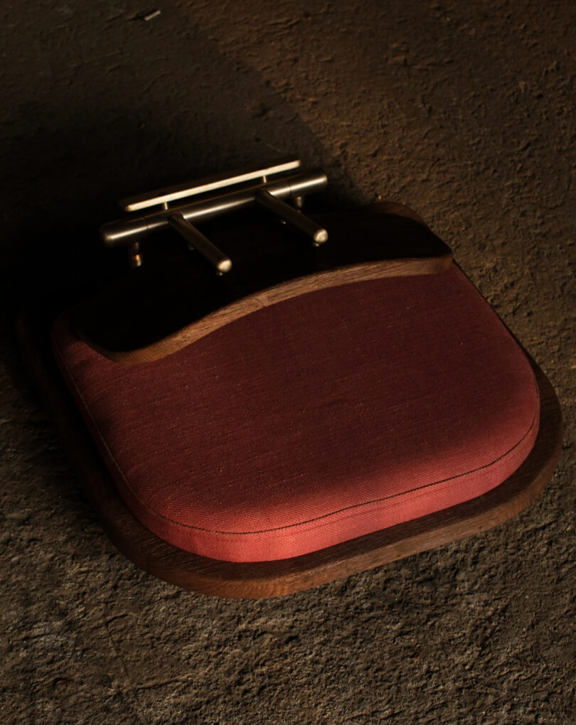 A maroon cushioned seat with dark wooden edges lies upside down on a textured brown surface, revealing metal hardware underneath.