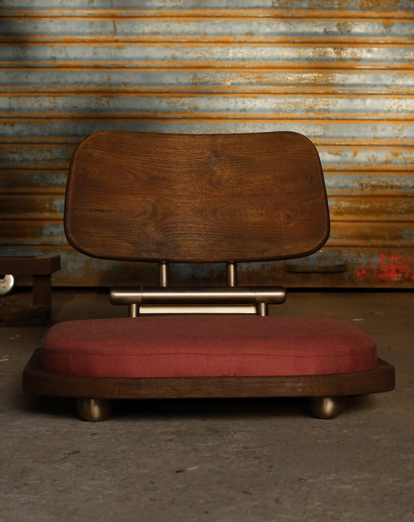 Low wooden chair with a curved backrest and red cushioned seat, placed on a concrete floor in front of a corrugated metal wall.