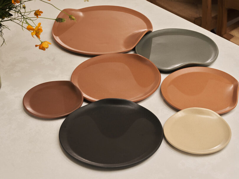 Seven ceramic plates of various sizes and earthy tones are arranged on a white tabletop beside a vase with yellow flowers.