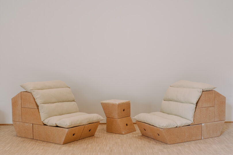 Two cushioned lounge chairs and a small matching side table made of light-colored wood are arranged on a wooden floor against a plain white wall