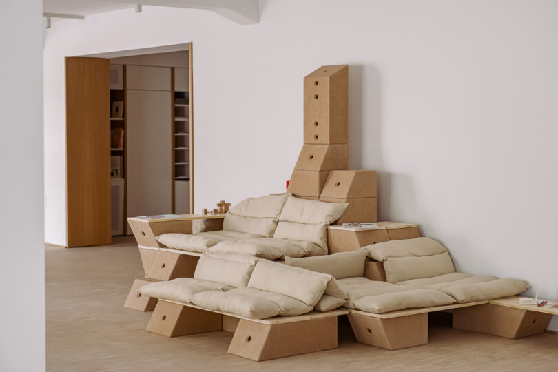 Modular furniture made of stacked cardboard and beige cushions is arranged against a white wall in a minimalist room with light wood flooring