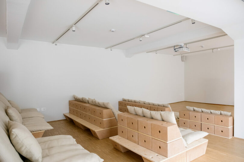 A minimalist room with modular, light brown block seating arranged in rows, each topped with white cushions, and a projector mounted on the ceiling