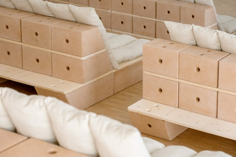 Modular wooden seating made from stacked rectangular blocks with white seat cushions and pillows in a minimalist indoor setting