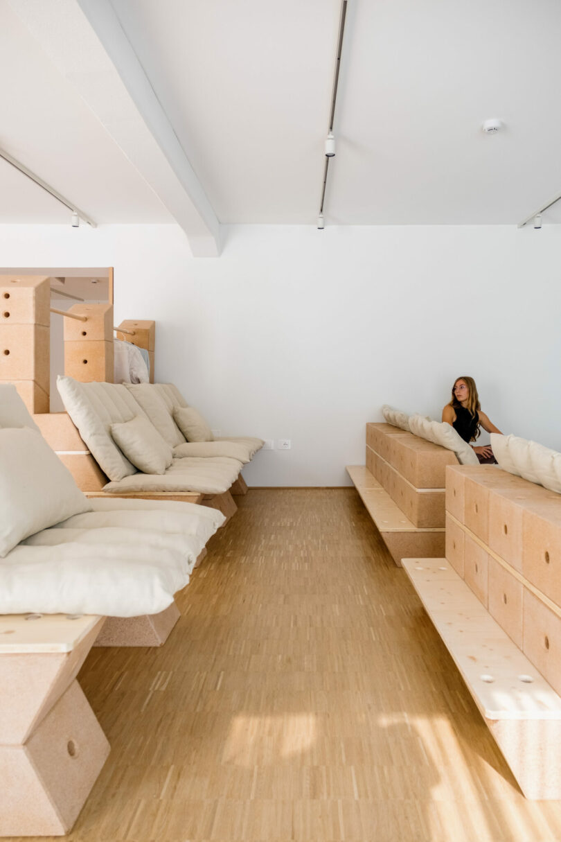A woman sits alone on a modern, minimalist wooden bench with white cushions in a bright, spacious room with light wood flooring