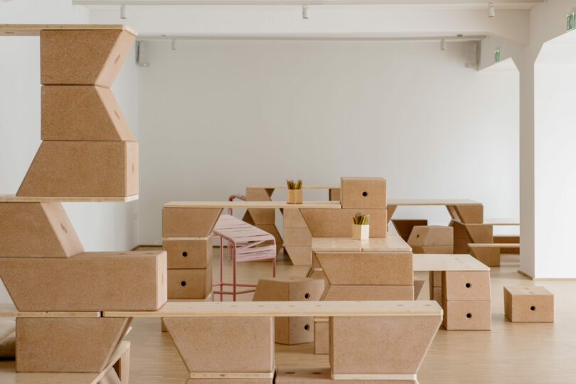 Minimalist room with modular wooden and cork furniture, including tables, benches, stools, and stacked blocks; some surfaces hold cups filled with pencils