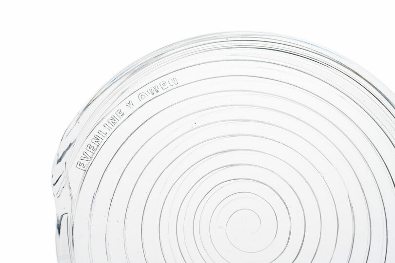A close-up of a clear glass plate with concentric circular ridges and embossed text reading "EVENLINE Y QWEEK" along the edge.