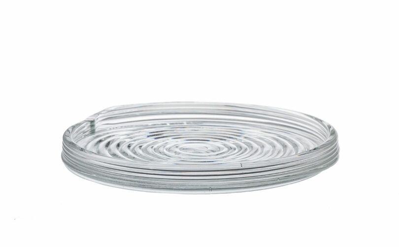A clear, round glass coaster with a ridged, spiral pattern, viewed from the side against a white background.
