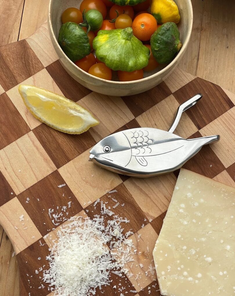 A stainless steel fish-shaped citrus squeezer on a checkered wooden board next to a bowl of veggies/tomatoes, a lime, some grated parmesian, and a block of parmesian.