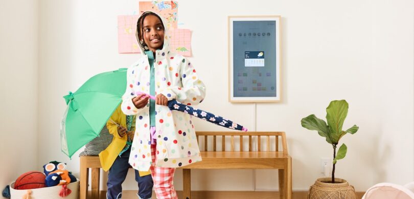 Two children in rain gear stand in a bright room; one holds a blue umbrella, the other holds a green umbrella near a wooden bench, with a digital calendar and plant on the wall