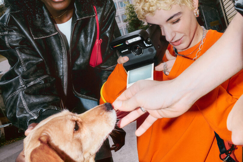 A person in an orange shirt uses an instant camera to photograph a dog being offered a treat by an outstretched hand, while another person stands nearby.