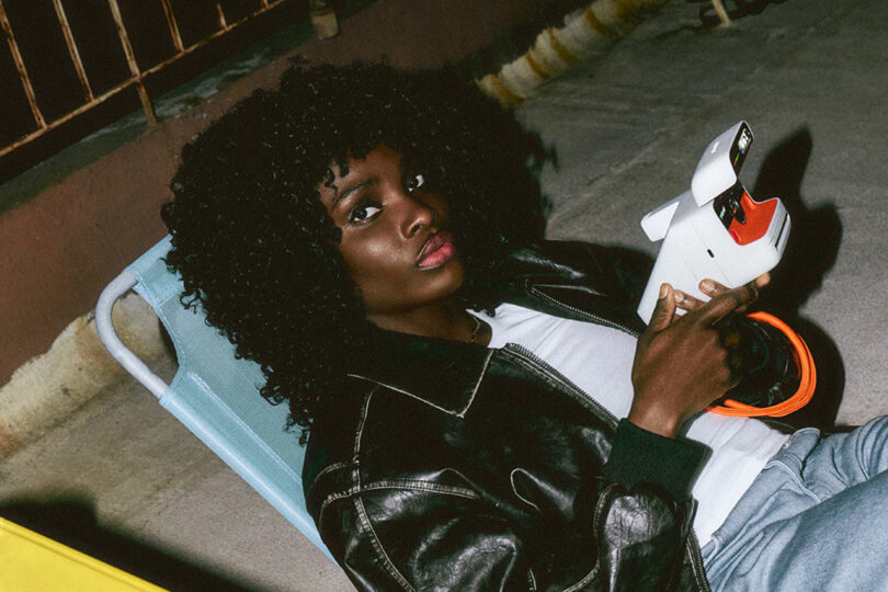 A person with curly hair and a black leather jacket sits on a chair holding an instant film camera, looking toward the camera, in an outdoor setting.