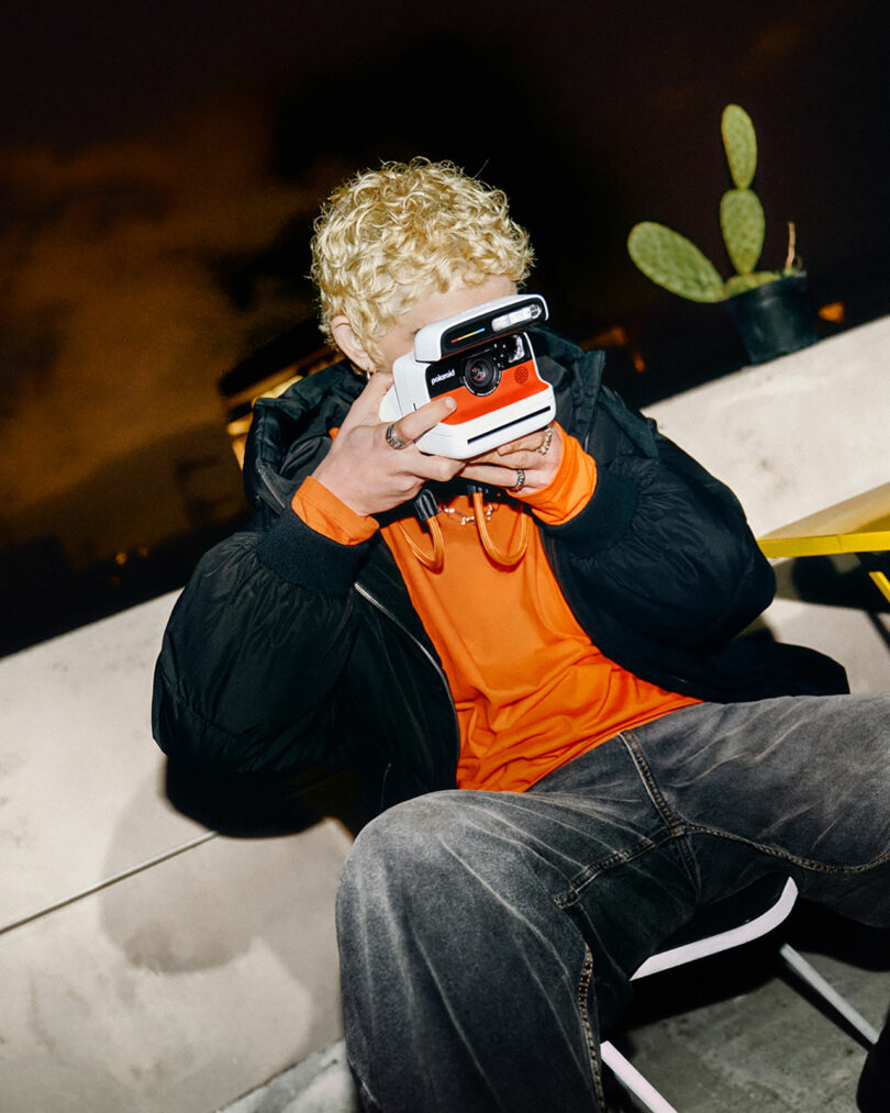 A person with curly blond hair, wearing an orange shirt and black jacket, sits on a chair outdoors at night, taking a photo with an instant camera. A potted cactus is visible in the background.