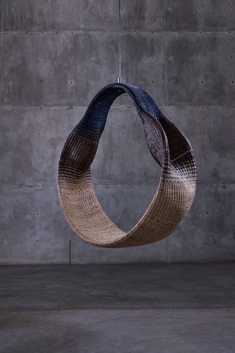 A woven chair hangs suspended from the ceiling in front of a gray concrete wall
