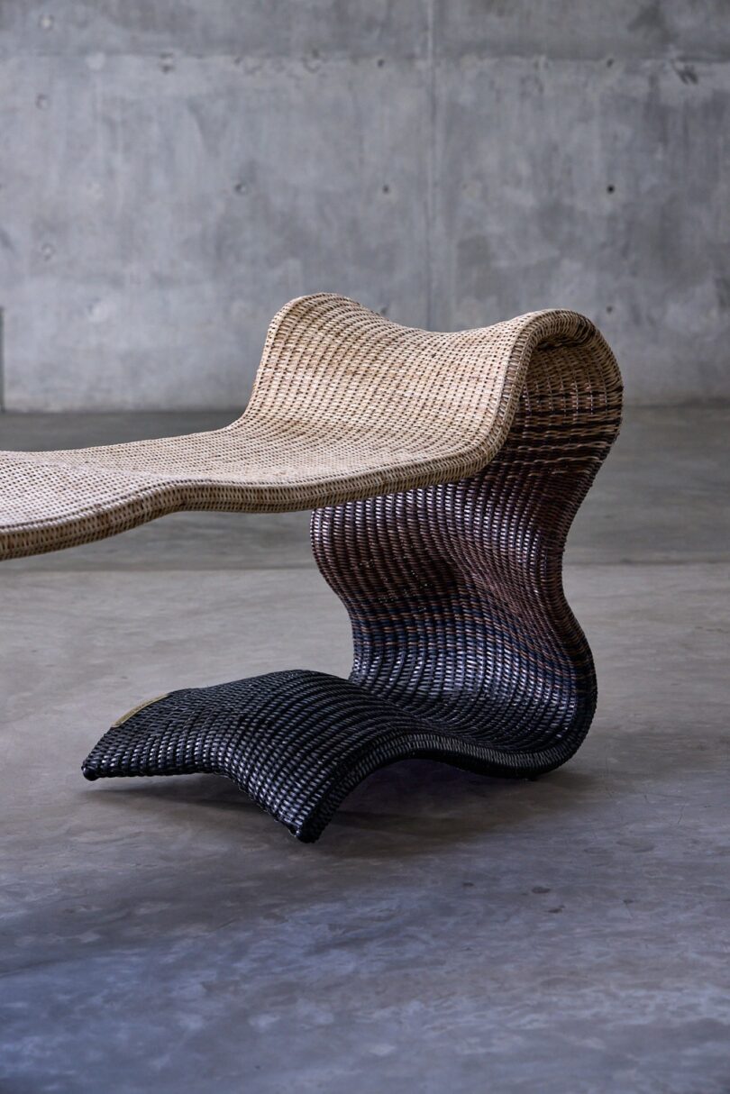 A modern, sculptural chair made of woven material in gradient shades from beige to dark brown, placed on a concrete floor with a gray textured wall in the background