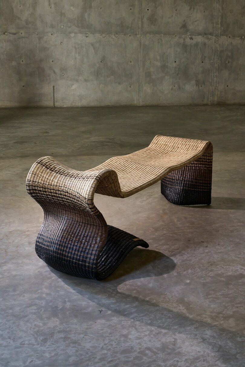 A woven, wave-shaped bench with a gradient from light tan to dark brown sits on a concrete floor against a bare concrete wall