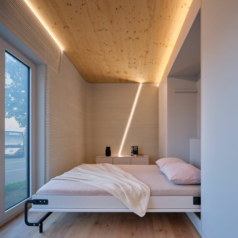 Minimalist bedroom with light wood ceiling, a neatly made bed with white bedding, a blanket, and a modern lamp on a small dresser near a large window.