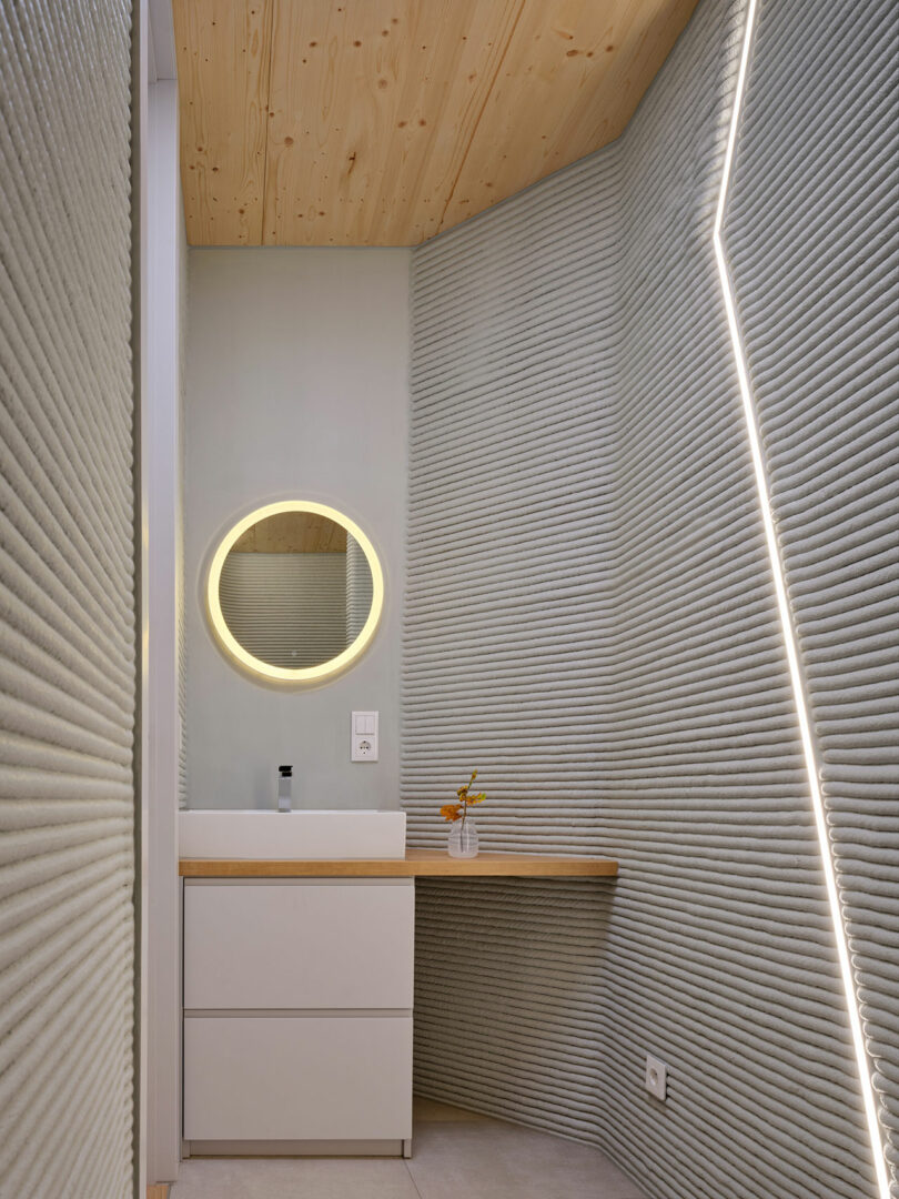 Modern bathroom with textured gray walls, a round backlit mirror above a white sink cabinet, a wooden countertop, and a geometric LED wall light.