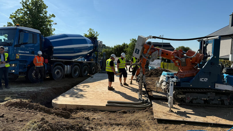 Workers in safety vests supervise a concrete mixer truck and a robotic machine pouring and leveling concrete at a construction site on a sunny day.
