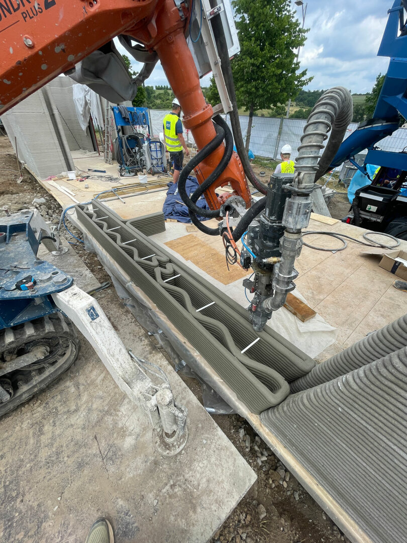 A large 3D printer extrudes concrete to construct a wall at an outdoor building site, with workers and construction equipment in the background.