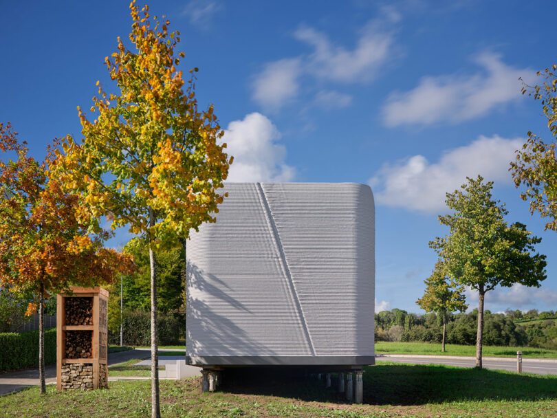 A small, modern 3D-printed concrete house stands elevated on short stilts in a grassy area, surrounded by trees with autumn foliage under a partly cloudy sky.