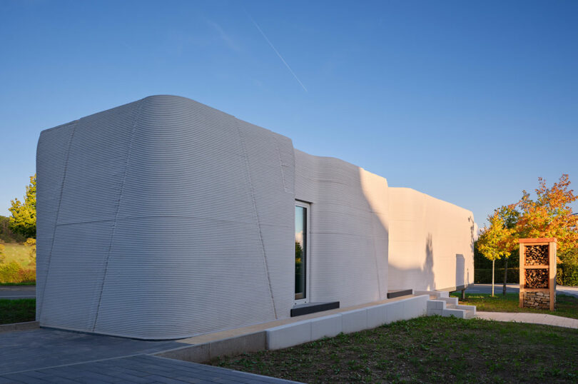 A modern, single-story building with curved, layered exterior walls, likely constructed using 3D printing technology, set against a clear blue sky with some trees nearby.