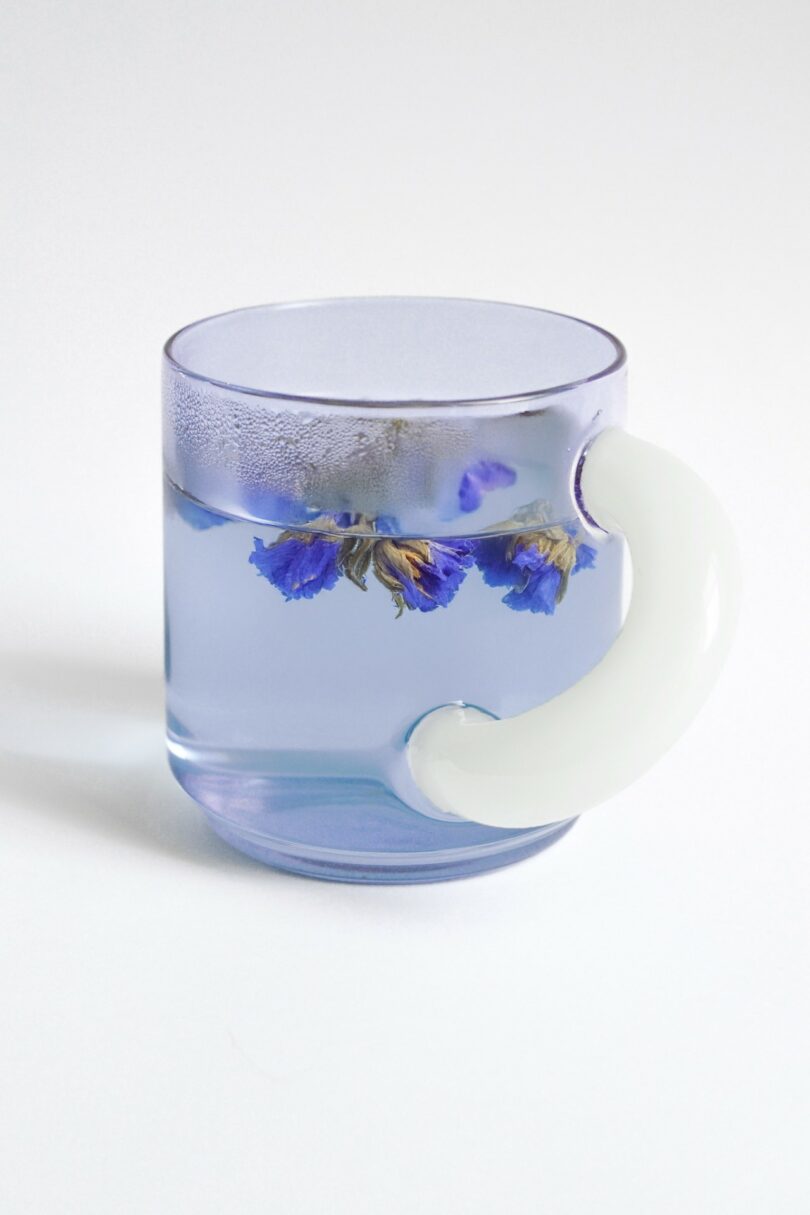 A transparent glass mug with a white handle, filled with clear liquid and floating purple flowers, placed on a white surface