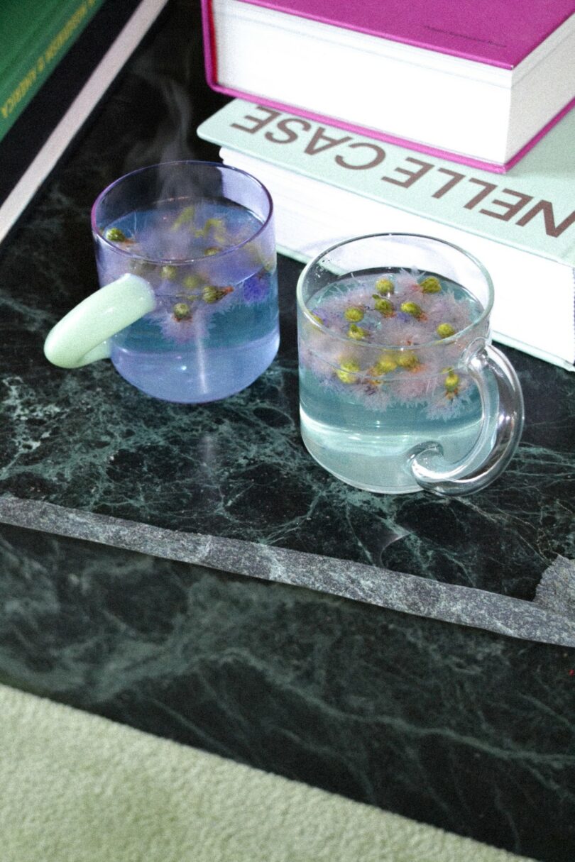 Two glass mugs filled with chamomile tea, with small flowers floating on top, placed on a dark marble table next to stacked books
